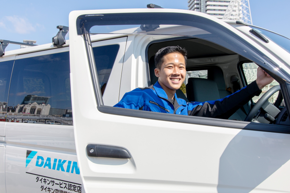 ダイキンのロゴが入った白い車の運転席から、青い作業着を着た男性が笑顔を見せている写真です。