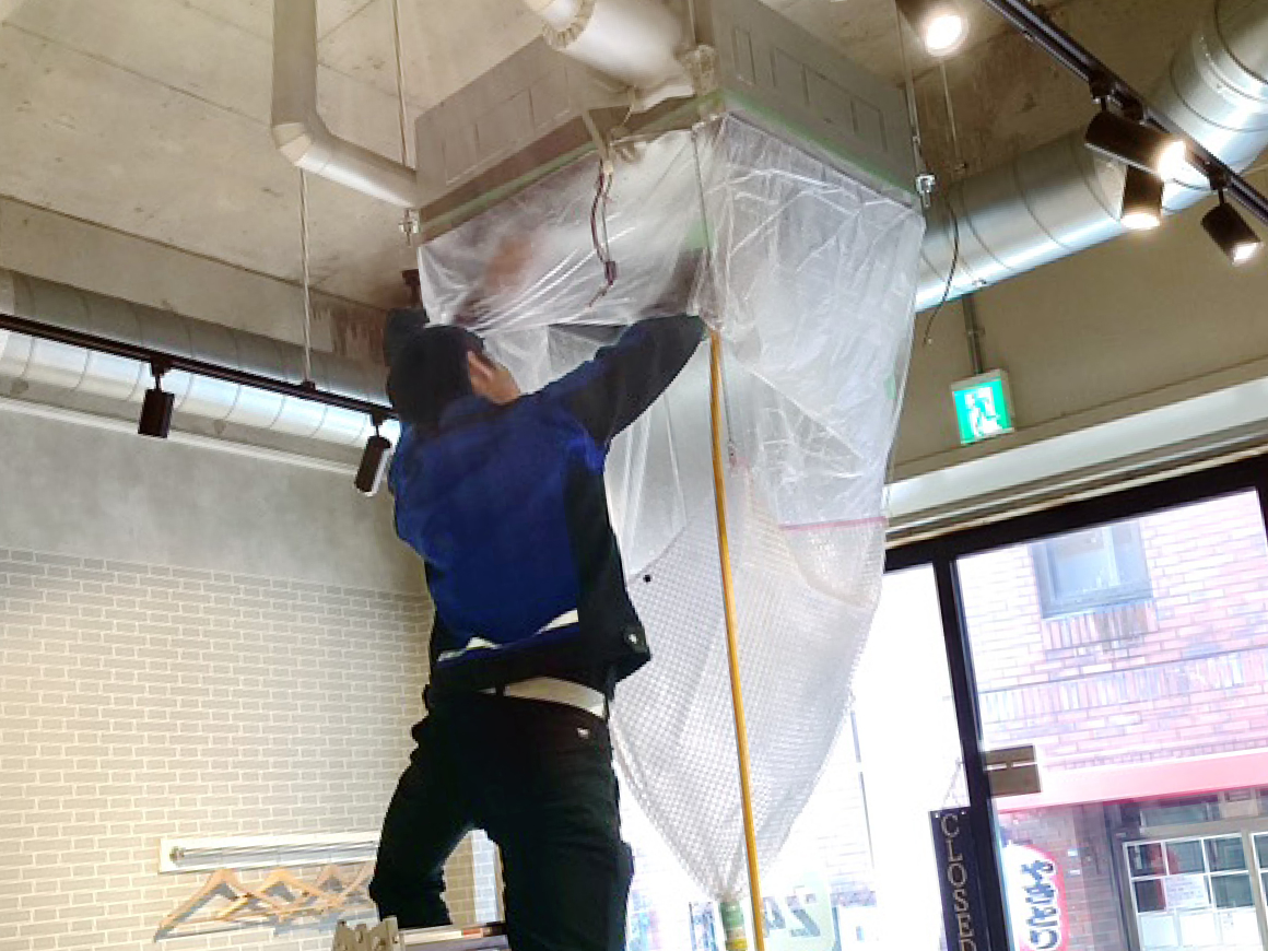 作業員が脚立に乗り、養生シートに覆われたエアコン内部の洗浄作業を行っている様子