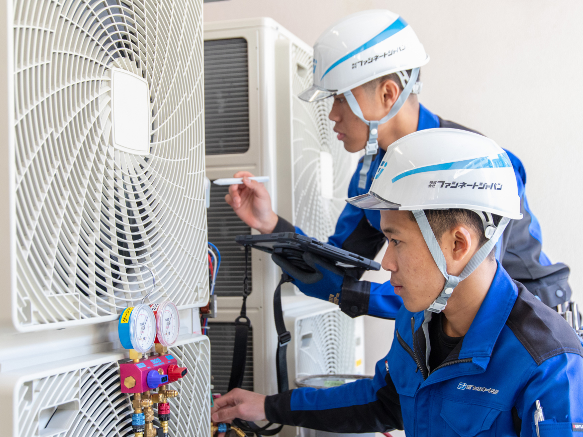 人の作業員が連携してエアコン室外機の点検を行っており、1人が計器を見ながら作業し、もう1人がタブレット端末で確認や記録をしている様子