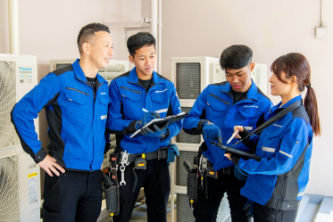 作業着姿の男女4名が、屋外の空調室外機の前でバインダーやタブレットを手に笑顔で打ち合わせをしている様子